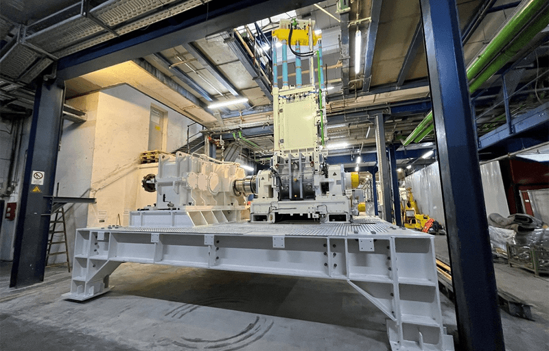 Large industrial machine mounted on a steel platform inside a factory hall, surrounded by beams, pipes and equipment in a spacious workshop setting.