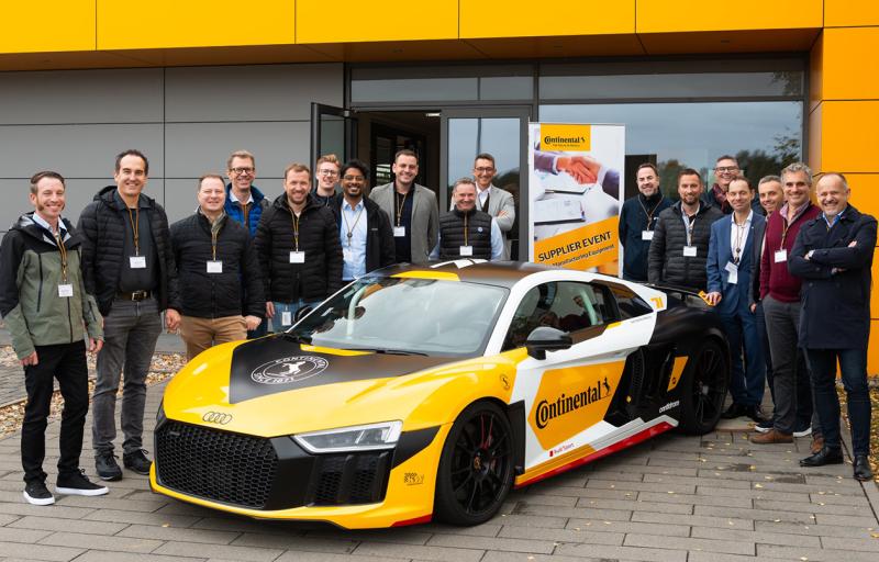 Group of people posing with a yellow and black Continental branded sports car outside a building during a supplier event.