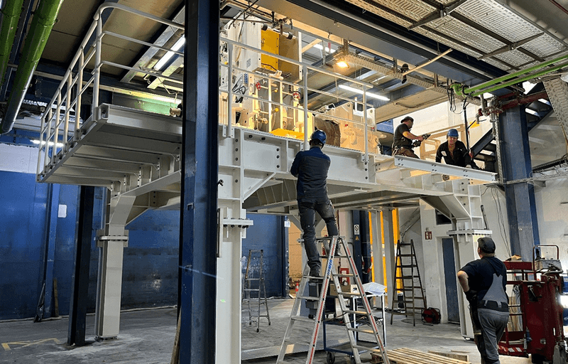 Workers install and service a large industrial machine on a raised platform in a factory hall, using ladders and tools amid steel beams and piping.
