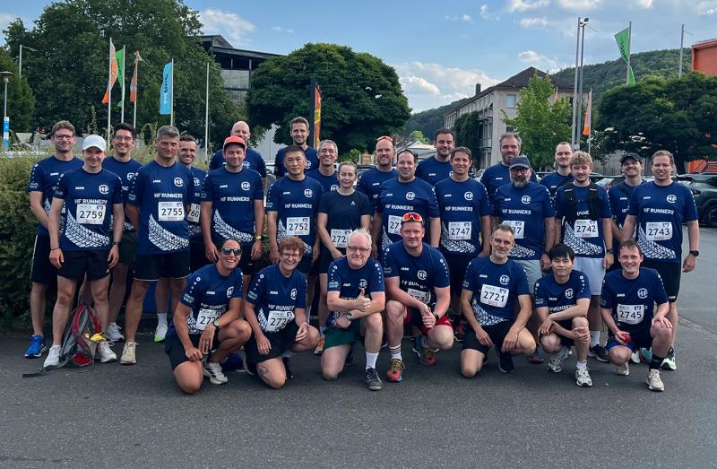 A group of runners in matching blue jerseys, each wearing race numbers, posing together on a street with trees and buildings in the background