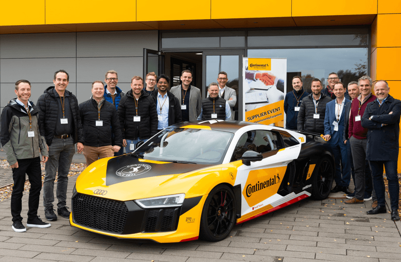 A group of people posing in front of a yellow and black Continental-branded sports car at a supplier event, with a banner visible in the background.