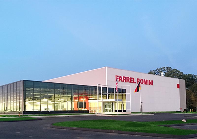 alt="Industrial building with "Farrel Pomini" sign, glass facade, and US and German flags."