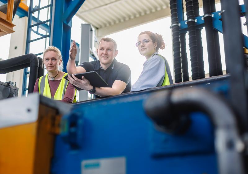 Three technicians in safety vests inspect equipment; one points with a tablet toward components, industrial setting with pipes and structures.