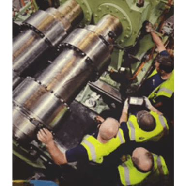Several technicians in safety vests work on a large rolling machine.