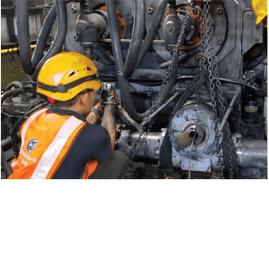 Technician wearing hard hat and safety vest works on an industrial machine; hoses, chains, and components visible during on-site maintenance.
