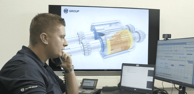 Service technician on phone in front of screen with 3D gearbox model; laptop and monitor display data for technical support and diagnostics.